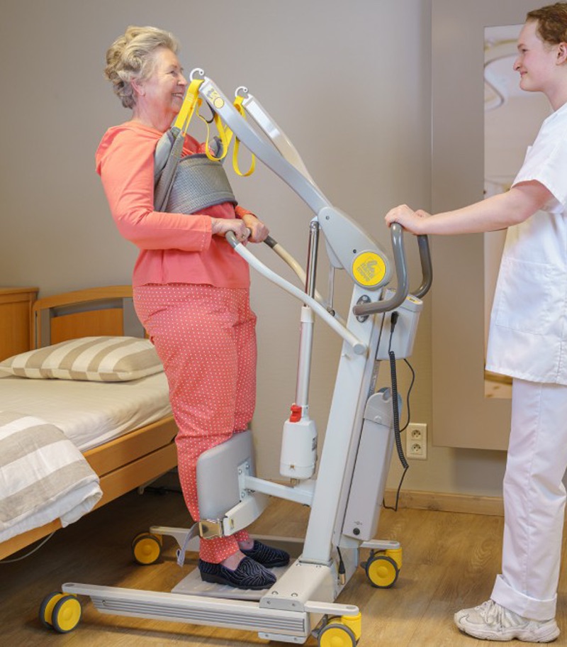 surehands-mobile-patient-lift-2620-06 SureHands 2620 Stand Assist Mobile Patient Lift providing standing support beside a bed, with a caregiver assisting a user during a safe mobility transfer in a healthcare setting.