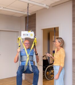 Caregiver operating a SureHands portable ceiling lift to safely transfer a seated individual using a supportive sling, demonstrating comfortable and secure patient lifting in an indoor care environment.