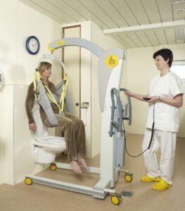 A caregiver operates a SureHands mobile patient lift using a handheld control to safely transfer a seated individual from a toilet in a home or care setting. The person is supported in a SureHands Body Support sling, while the portable lift provides stable, powered lifting for comfortable bathroom transfers.