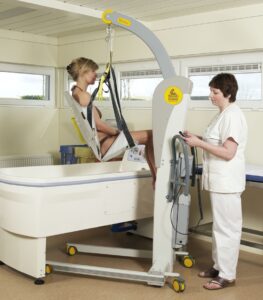 A caregiver uses a SureHands Victor 2600 mobile patient lift with a supportive bathing sling to safely transfer an individual into a therapeutic bathtub. The portable powered lift provides stable, comfortable assistance for bathing and personal care in a healthcare or home care environment.