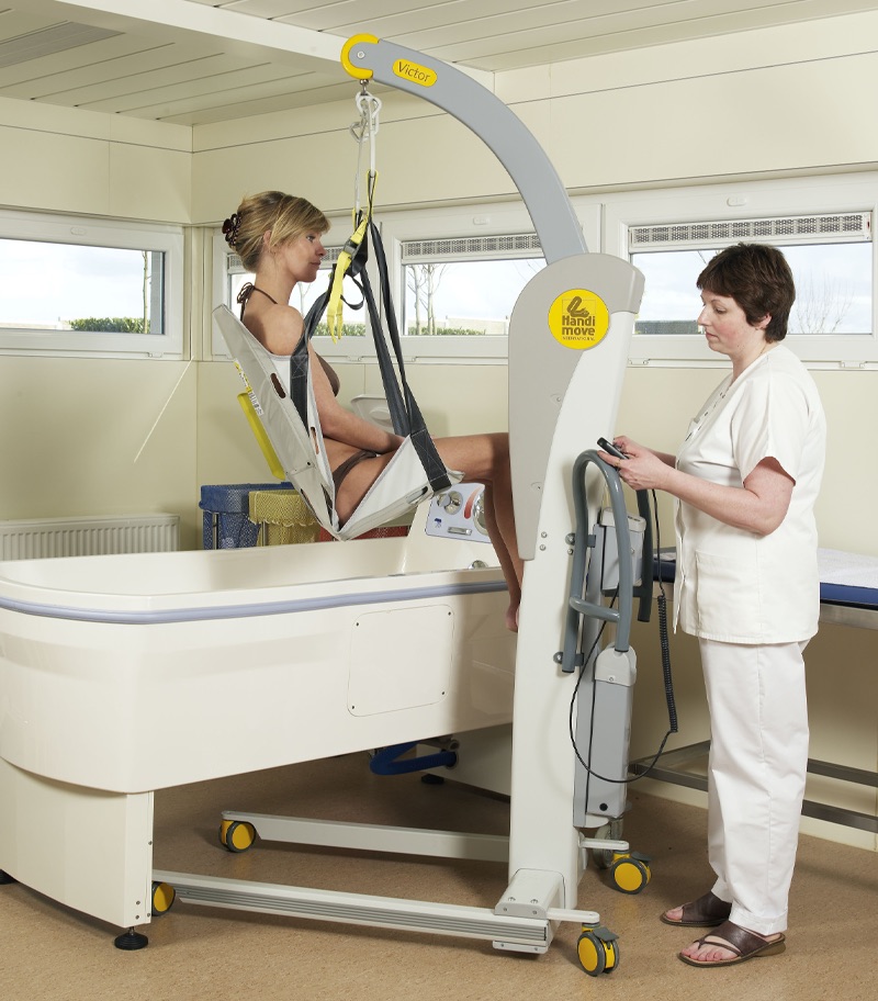 victor(2600)-mobile-patient-lift-bath-surehands A caregiver uses a SureHands Victor 2600 mobile patient lift with a supportive bathing sling to safely transfer an individual into a therapeutic bathtub. The portable powered lift provides stable, comfortable assistance for bathing and personal care in a healthcare or home care environment.