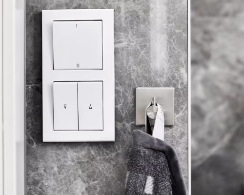 A close-up of the Accessible Lighting switch panel, designed for wheelchair access, mounted on a gray marble wall with a stainless steel hook nearby holding a dark towel with white patterns.